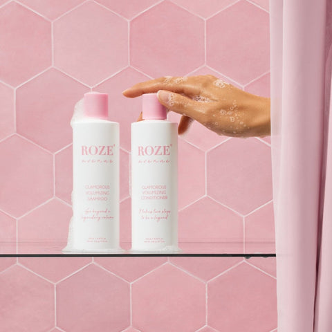 szampon i odżywyka Roze Avenue - glamorous volumizing shampoo i conditioner ustawione obok siebie na szklanej półce, ręka kobiety spod prysznica sięga po odżywkę